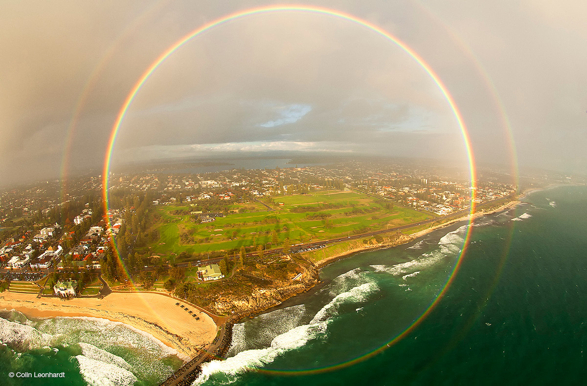 Circular double rainbow above sea, shore, paddocks and houses, with a grey, cloudy sky